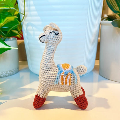 A small crocheted llama figurine with a cream body, red hooves, and a blue and yellow saddle, displayed on a white surface with potted plants in the background.