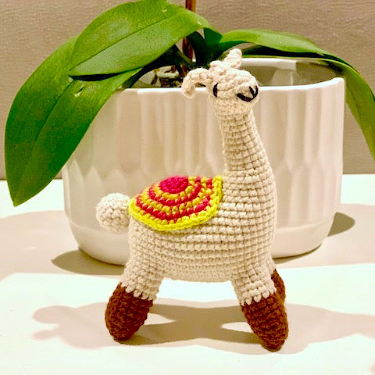 Crocheted llama toy with a striped, oval-shaped saddle in pink and chartreuse colors. 