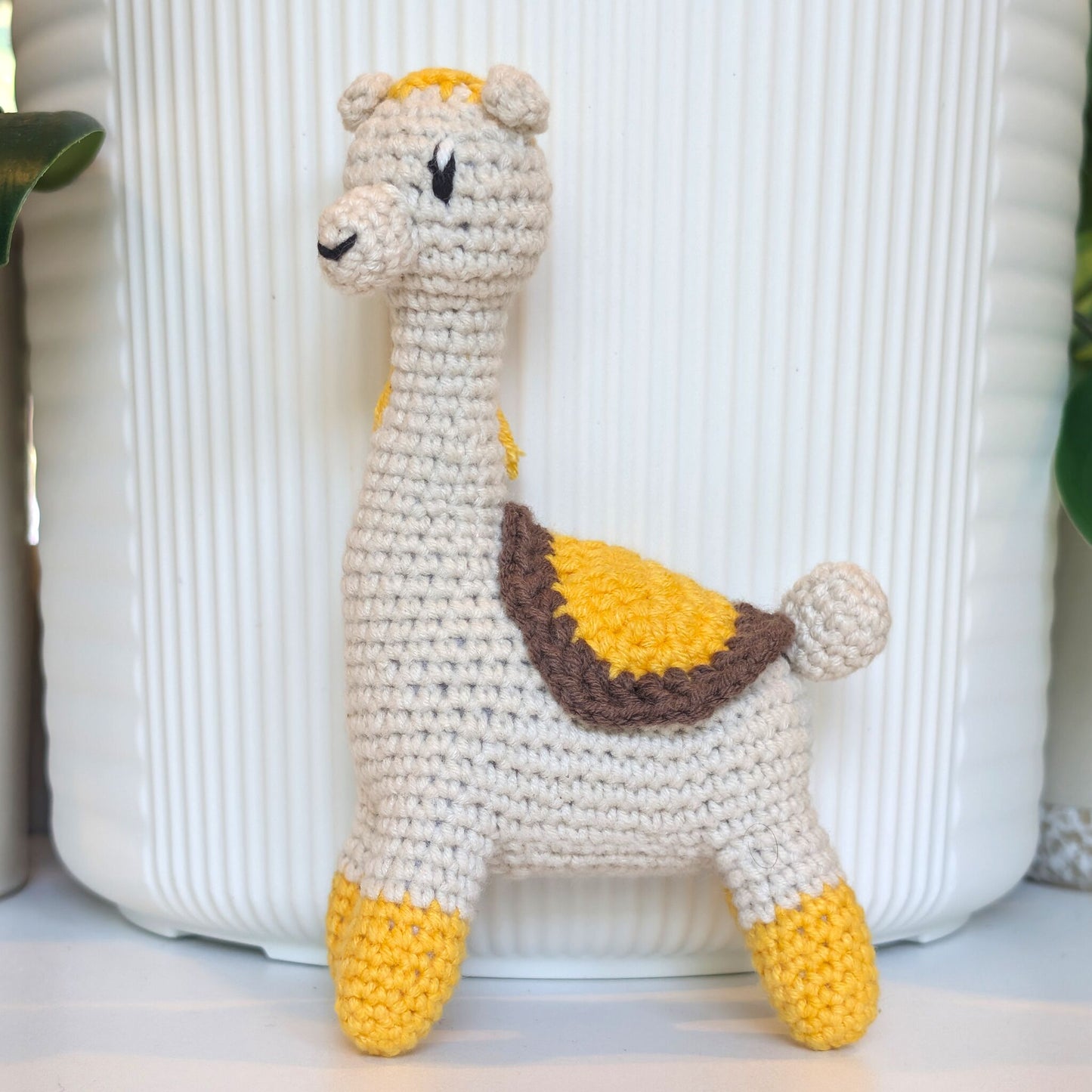 A handmade crocheted llama toy with a cream body, yellow hooves and crown, and a brown saddle, standing in front of a white ribbed ceramic pot.
