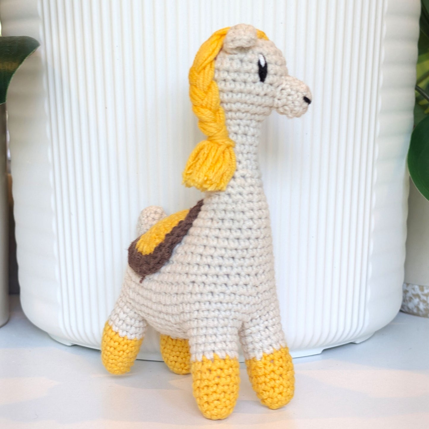 A crocheted llama plush toy with a cream body, yellow braided mane and hooves, and a brown saddle detail, standing in front of a white ribbed ceramic planter.