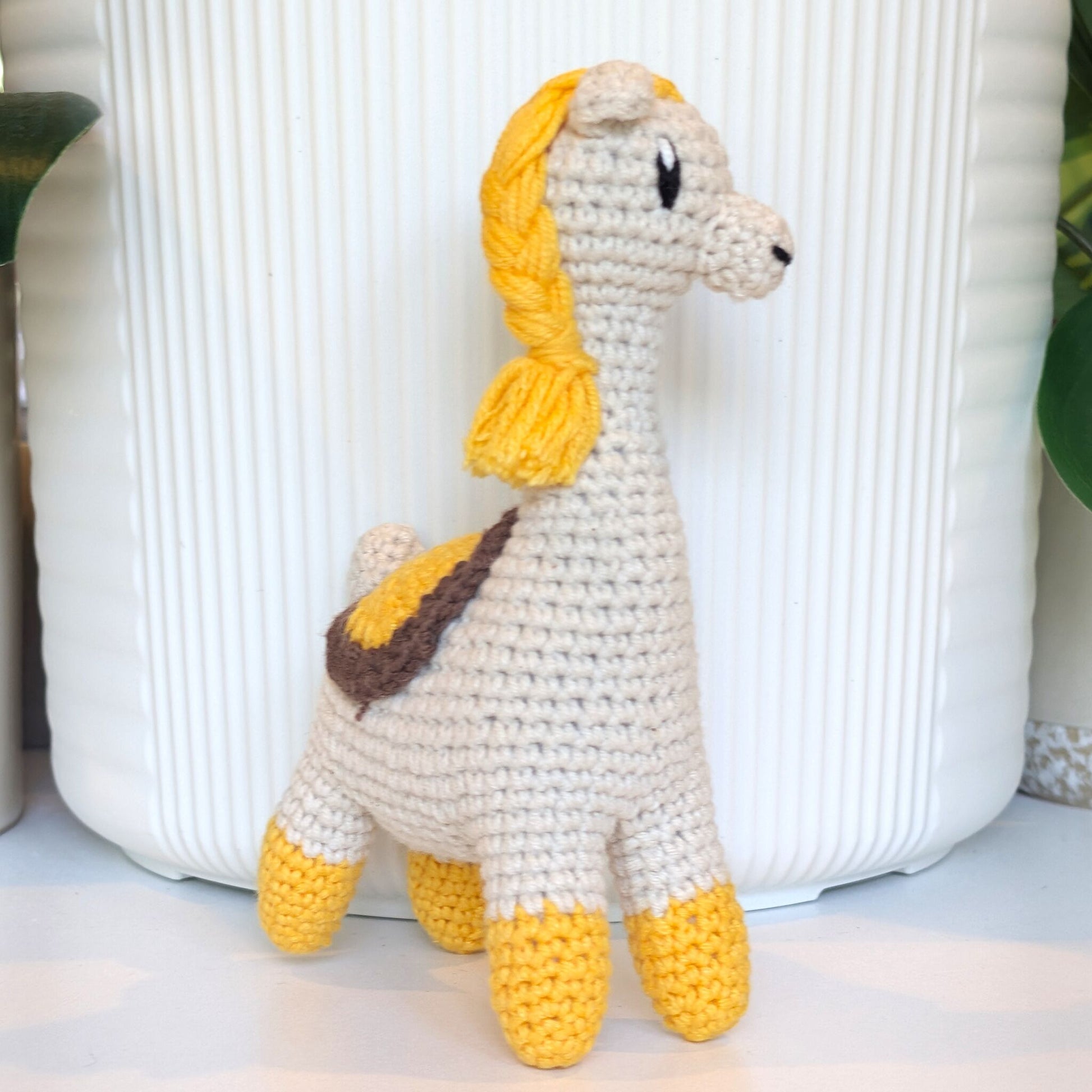 A crocheted llama plush toy with a cream body, yellow braided mane and hooves, and a brown saddle detail, standing in front of a white ribbed ceramic planter.