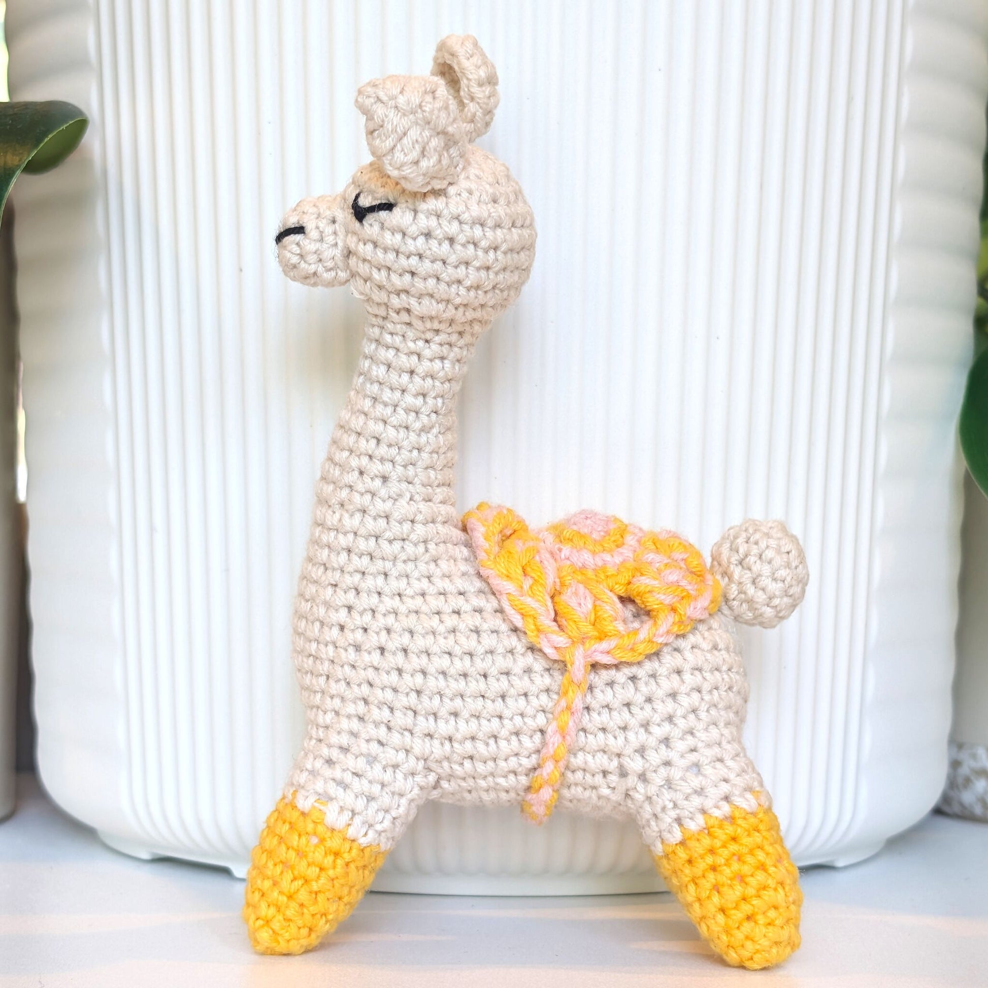 A handmade crocheted llama plush toy in cream with yellow and pink accents, standing in front of a white ribbed ceramic pot.