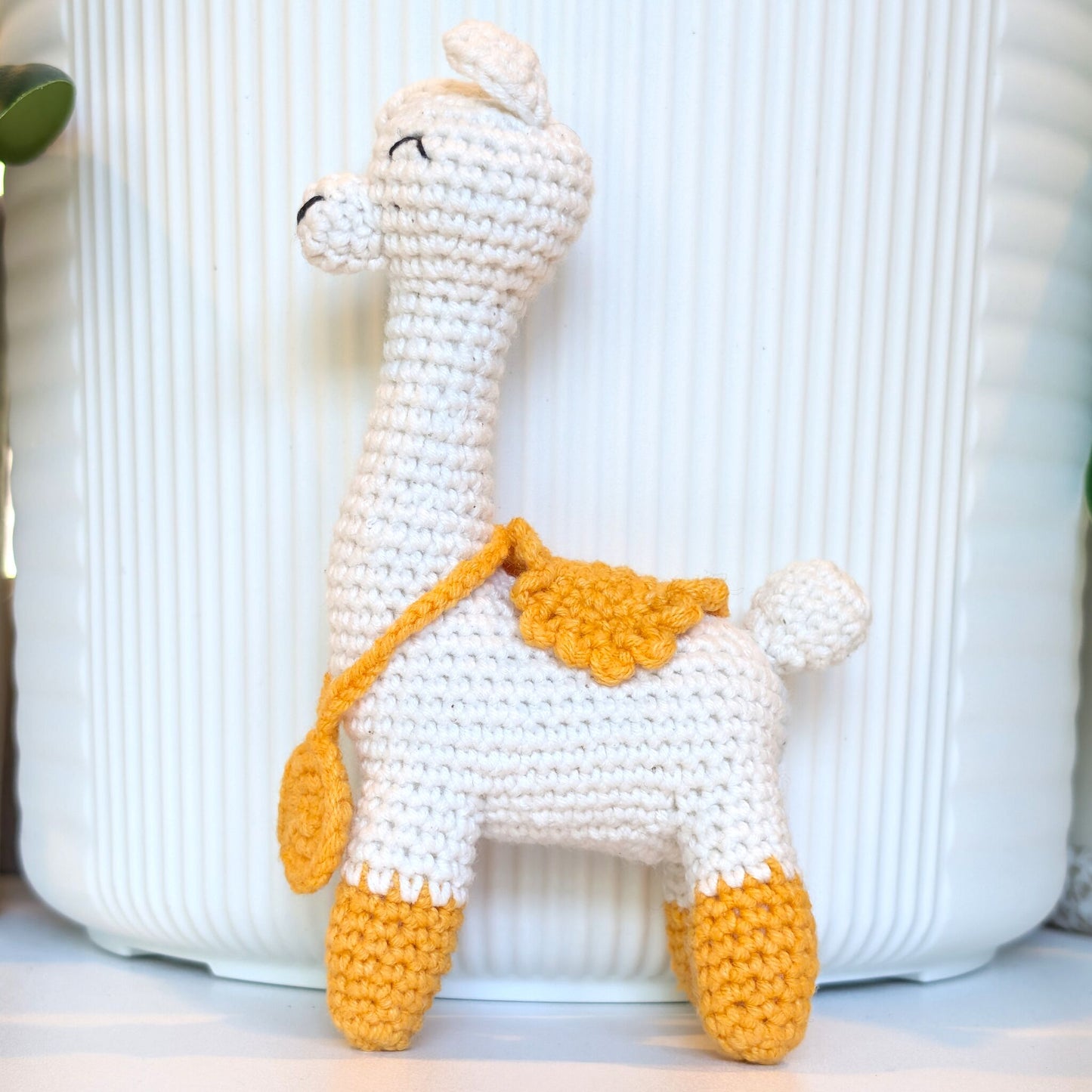 A handmade crocheted llama toy in off-white with mustard yellow accents on its hooves, saddle, and strap, standing in front of a white ribbed ceramic pot.