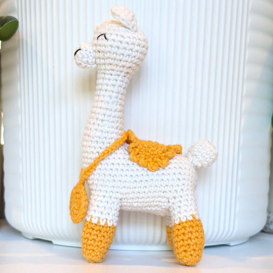 A handmade crocheted llama toy in off-white with mustard yellow accents on its hooves, saddle, and strap, standing in front of a white ribbed ceramic pot.