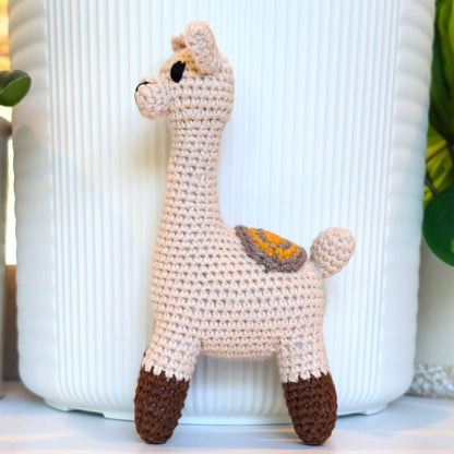 A crocheted llama toy standing in front of a white ribbed ceramic pot, with green leaves visible on the sides.