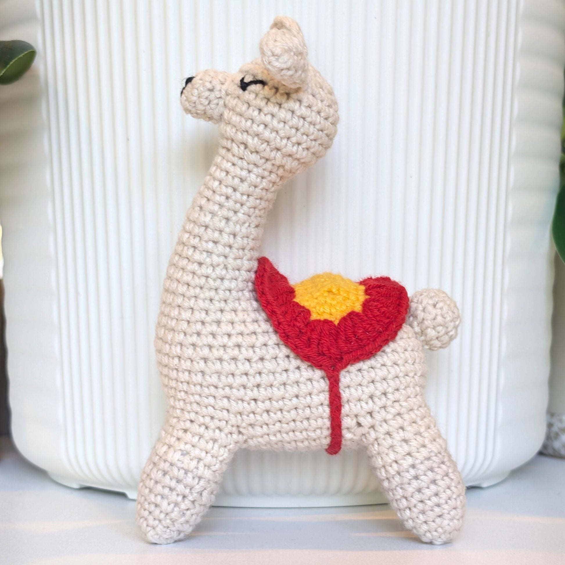 A handmade crocheted llama toy in cream-colored yarn, featuring a red and yellow flower patch on its back, set against a white ribbed ceramic background.