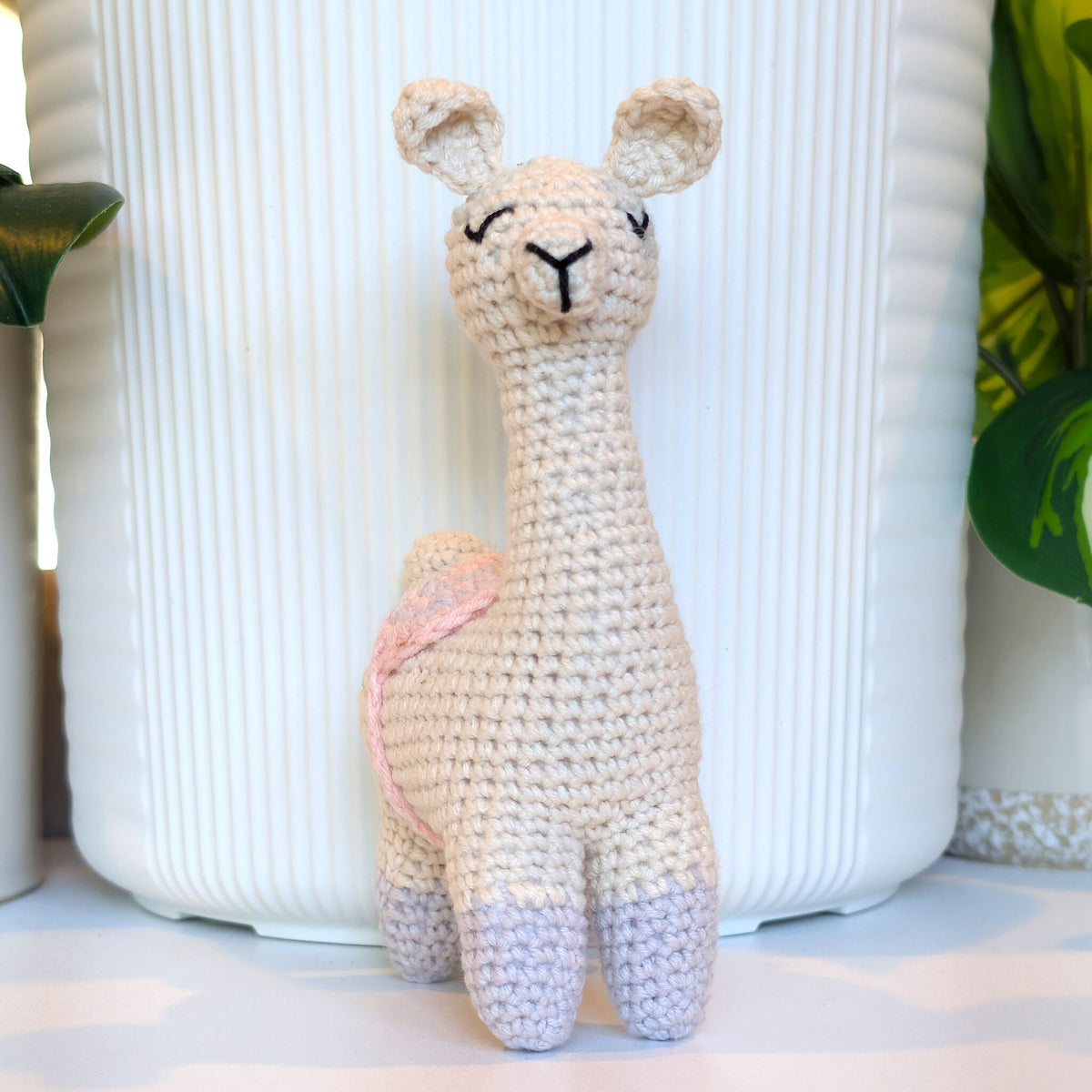 A handmade crocheted llama toy with gray boots and gray, pink saddle standing in front of a white ribbed ceramic vase.