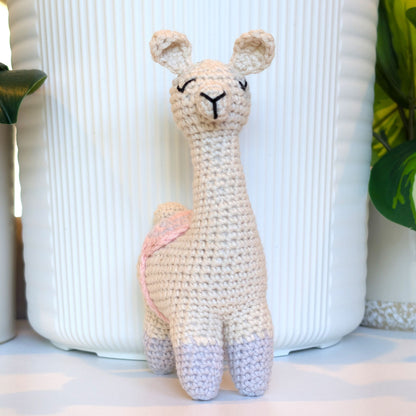A handmade crocheted llama toy with gray boots and gray, pink saddle standing in front of a white ribbed ceramic vase.