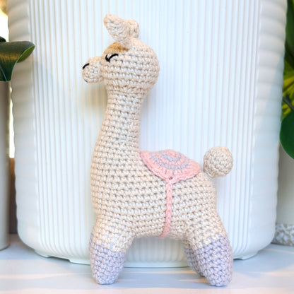 A handmade crocheted llama toy with gray boots and gray, pink saddle standing in front of a white ribbed ceramic vase.