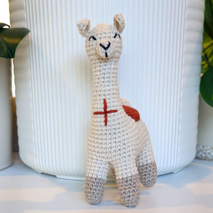 A handmade crocheted llama toy in off-white with a terracotta saddle on its back and gray feet, standing in front of a white ribbed ceramic vase.