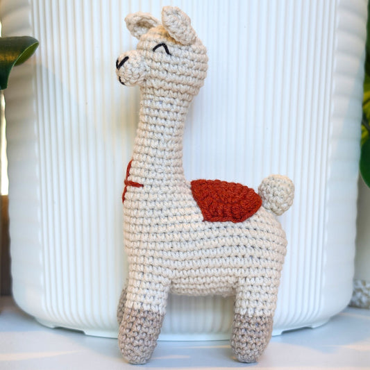A handmade crocheted llama toy standing in front of a white ribbed ceramic pot. The toy is cream-colored with a rust-red saddle and grey-painted hooves.