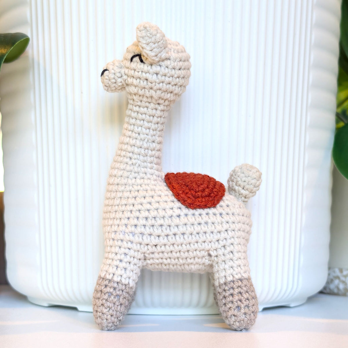 A handmade crocheted llama toy in off-white with a terracotta saddle on its back and gray feet, standing in front of a white ribbed ceramic vase.