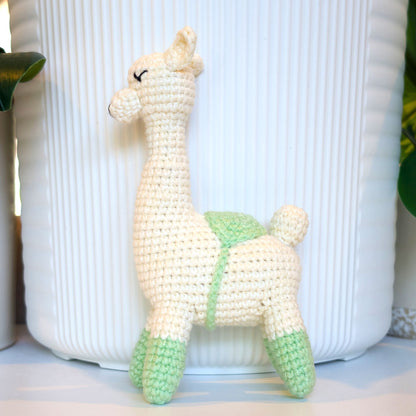 A handmade crocheted llama toy in cream and light mint green, standing in front of a white ribbed ceramic pot.