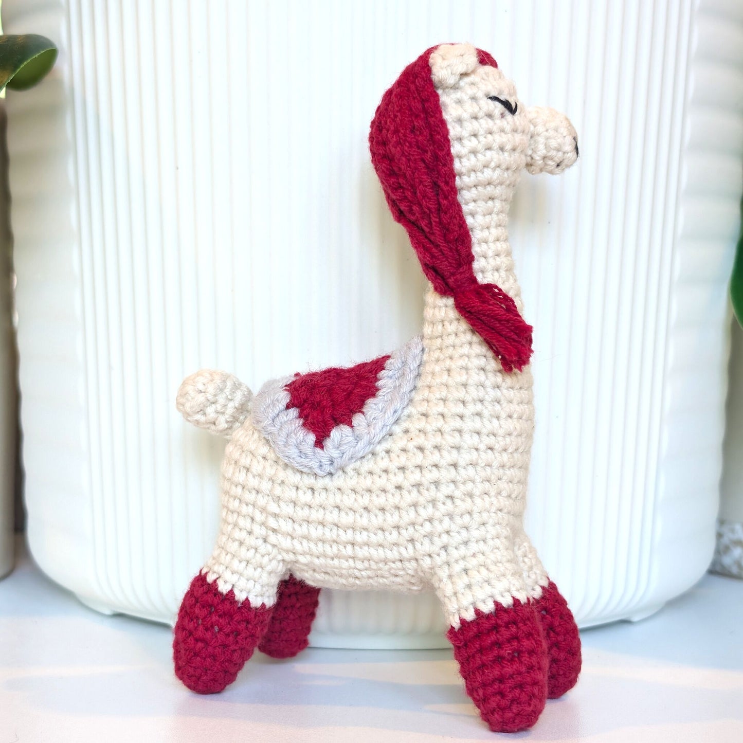 A handmade crocheted llama or alpaca doll in cream with red accents, standing in front of a white ribbed ceramic pot.