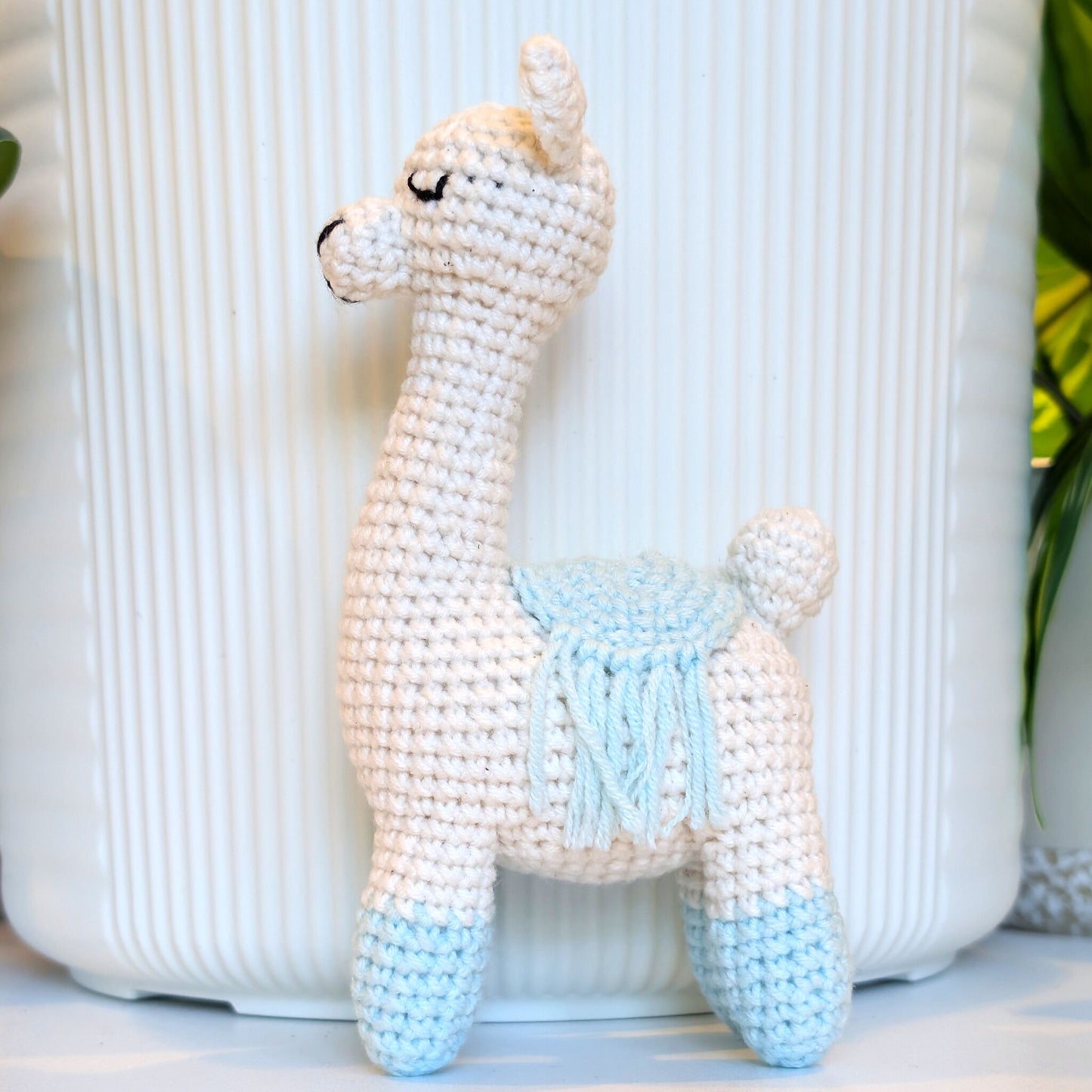 A crocheted llama toy with a cream body, light blue or pastel aqua accents on the legs and saddle, and black embroidered eyes, standing in front of a white ribbed container.