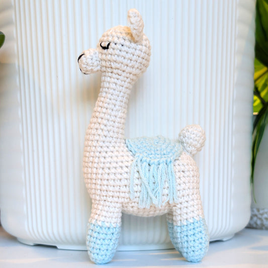 A crocheted llama toy with a cream body, light blue or pastel aqua accents on the legs and saddle, and black embroidered eyes, standing in front of a white ribbed container.