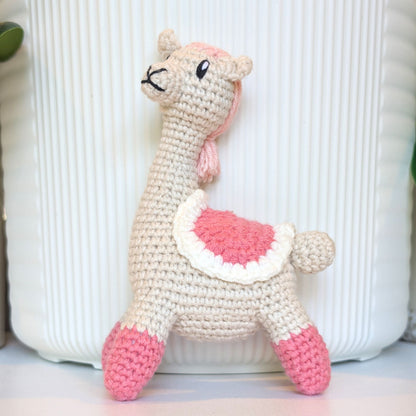 A handmade crocheted llama toy in cream, pink, and white saddle and long hair, standing in front of a white ribbed container.