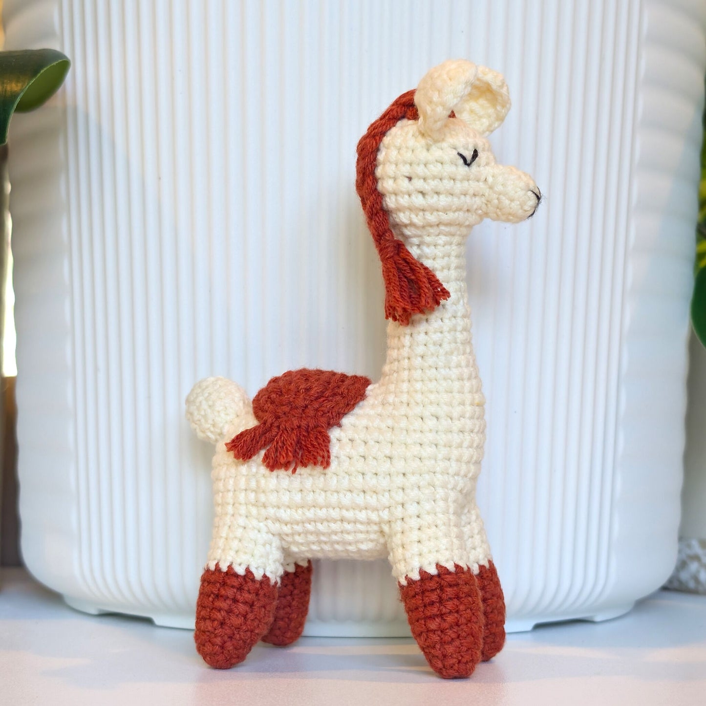 A crocheted llama or alpaca toy figure with a cream body and terracotta accents on the mane, back, and hooves, standing in front of a white ribbed ceramic pot.