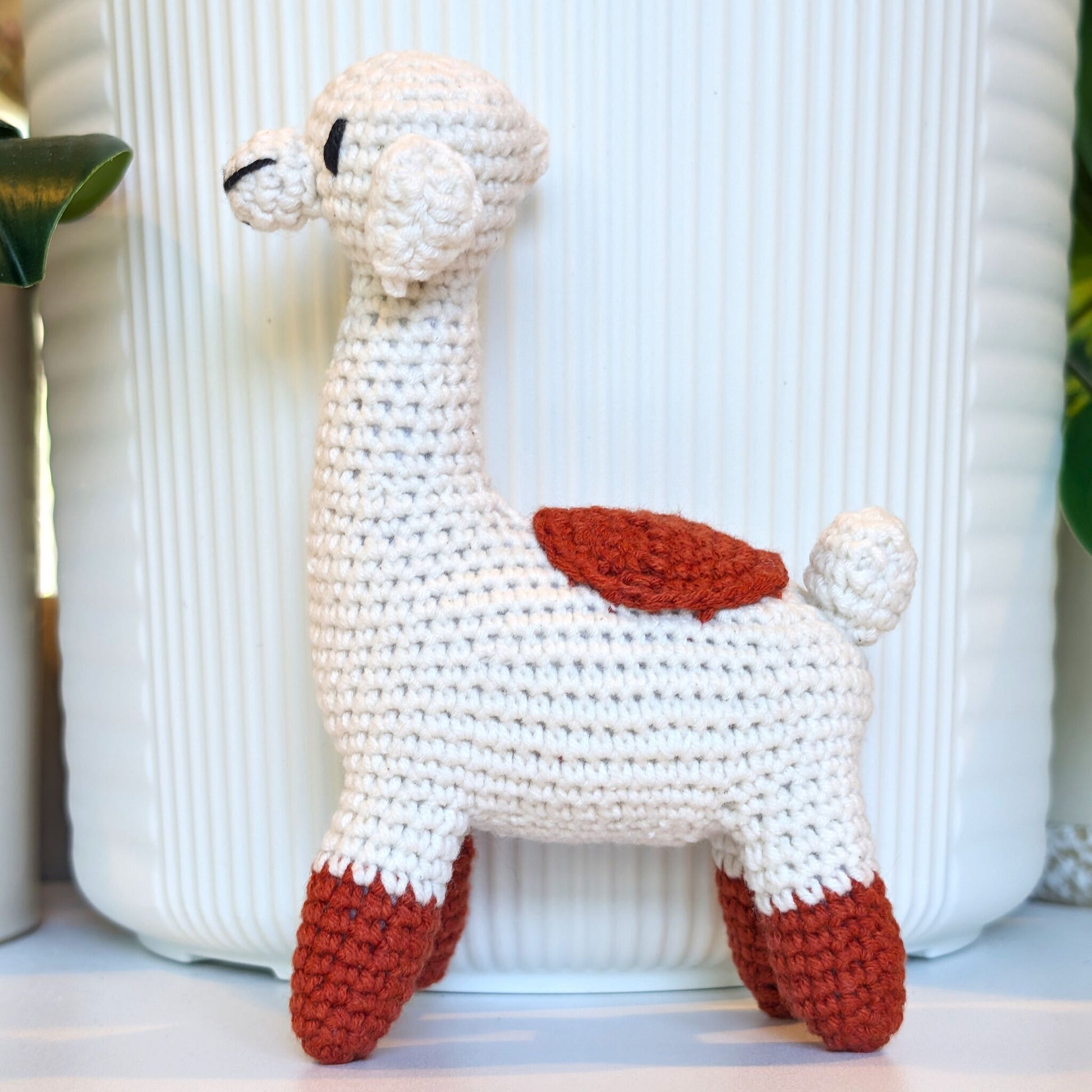 A handmade crocheted llama toy in off-white yarn with terracotta accents on its back and feet, standing in front of a white ribbed ceramic pot.