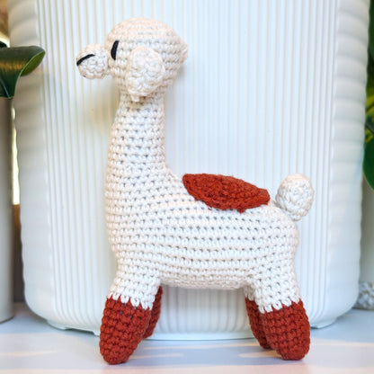 A handmade crocheted llama toy in off-white yarn with terracotta accents on its back and feet, standing in front of a white ribbed ceramic pot.