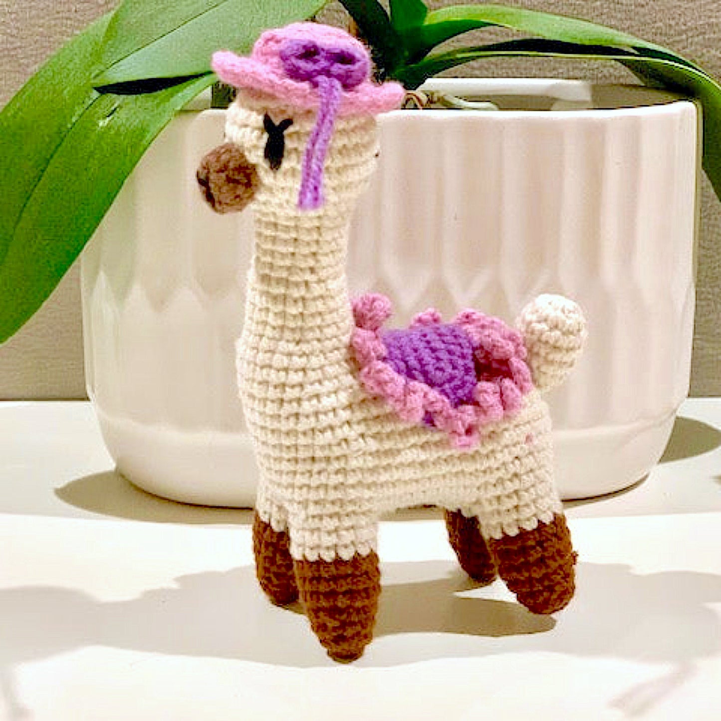 Crocheted llama toy with pink and purple accents in front of a white pot with green leaves.