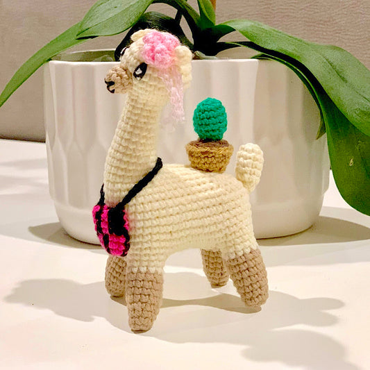 Crocheted plush llama toy with a cactus on its back and a pink bow and satchel