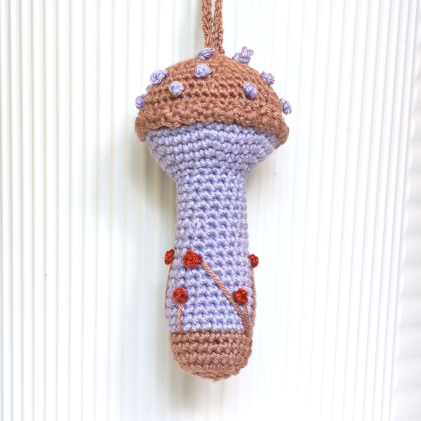 A handmade crocheted mushroom-shaped ornament or hanging decoration, featuring a light lavender body, brown cap and base, and small purple and red yarn accents, shown against a white ribbed background.