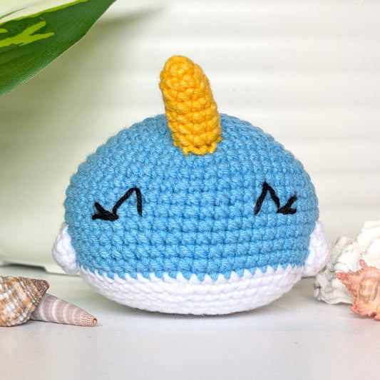 A crocheted narwhal plush toy with a light blue body, white underside, and a yellow horn, styled on a white surface with seashells and a green leaf in the background.