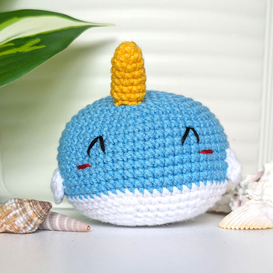 A crocheted narwhal plush toy with a light blue body, white underside, and a yellow horn, with squinting smiling embroidered eyes .Unisex.