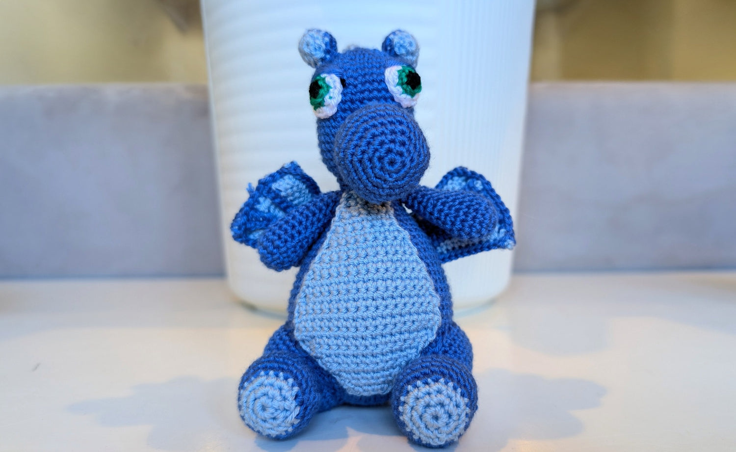 Refugee-made blue crocheted hippo toy sitting on a surface with a white cup in the background.