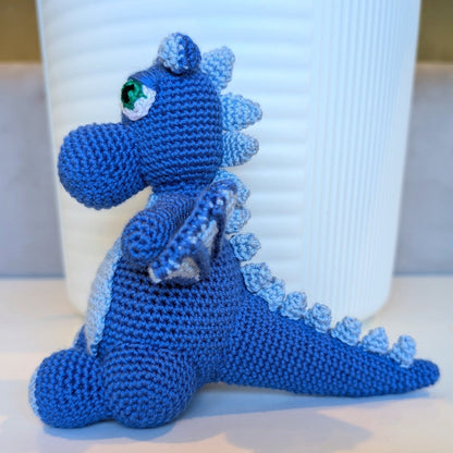 Refugee-made blue crocheted dragon toy in front of a white container on a light surface.