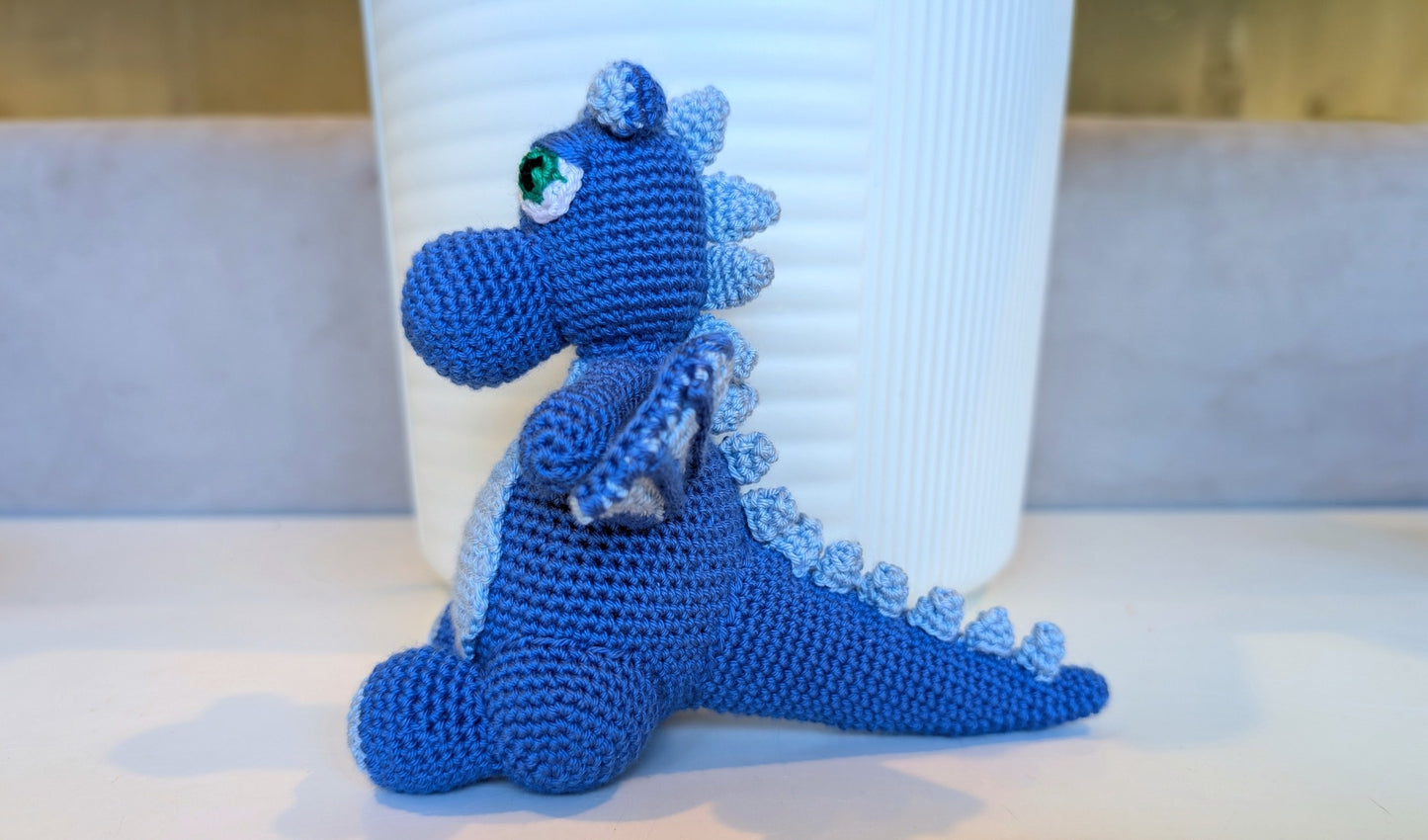 Refugee-made blue crocheted dragon toy in front of a white container on a light surface.