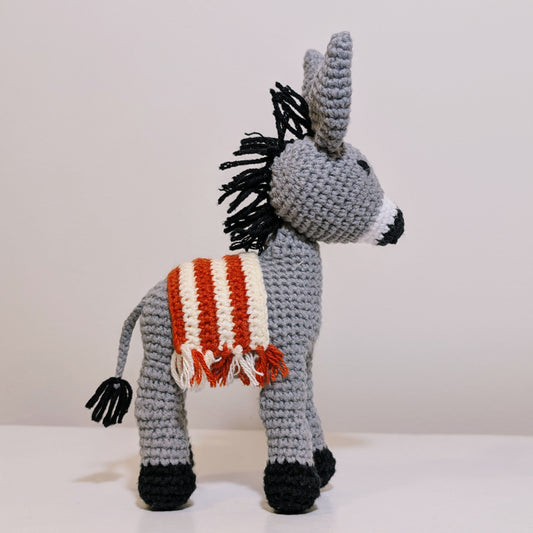 Crocheted plush donkey toy with a red-orange and cream fringed saddle in amigurumi style 