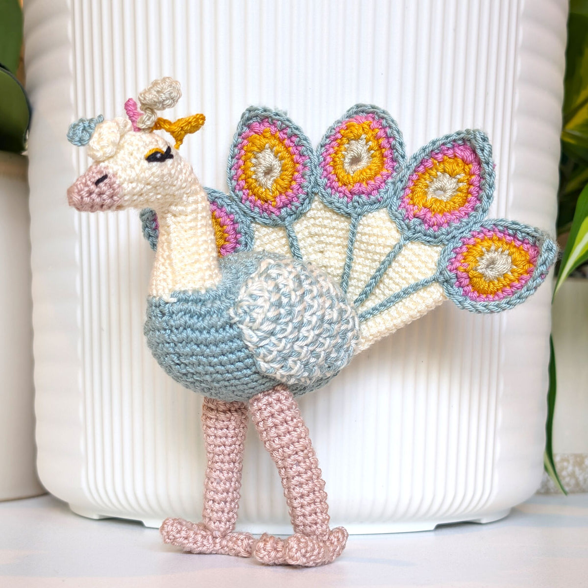 A crocheted peacock figurine with cream, light blue, pink, and yellow yarn, standing in front of a white ribbed ceramic pot.
