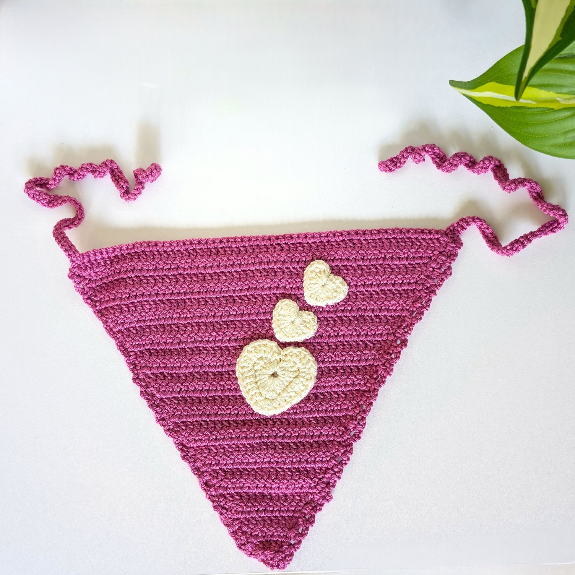 A handmade crochet triangle bikini top in magenta pink with three cream-colored heart appliqués on the front. The top has wavy shoulder straps and is photographed flat on a white surface with a green plant leaf in the upper right corner.