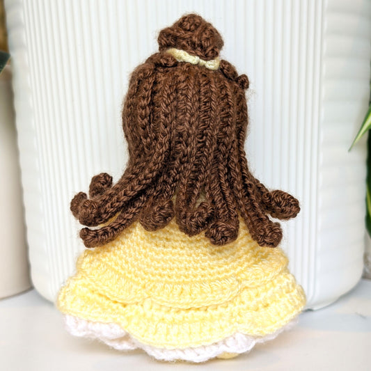 A hand-crocheted doll viewed from the back, featuring long brown yarn hair with a small cream ribbon and a yellow ruffled dress with a white lace hem, set against a white ribbed ceramic background.