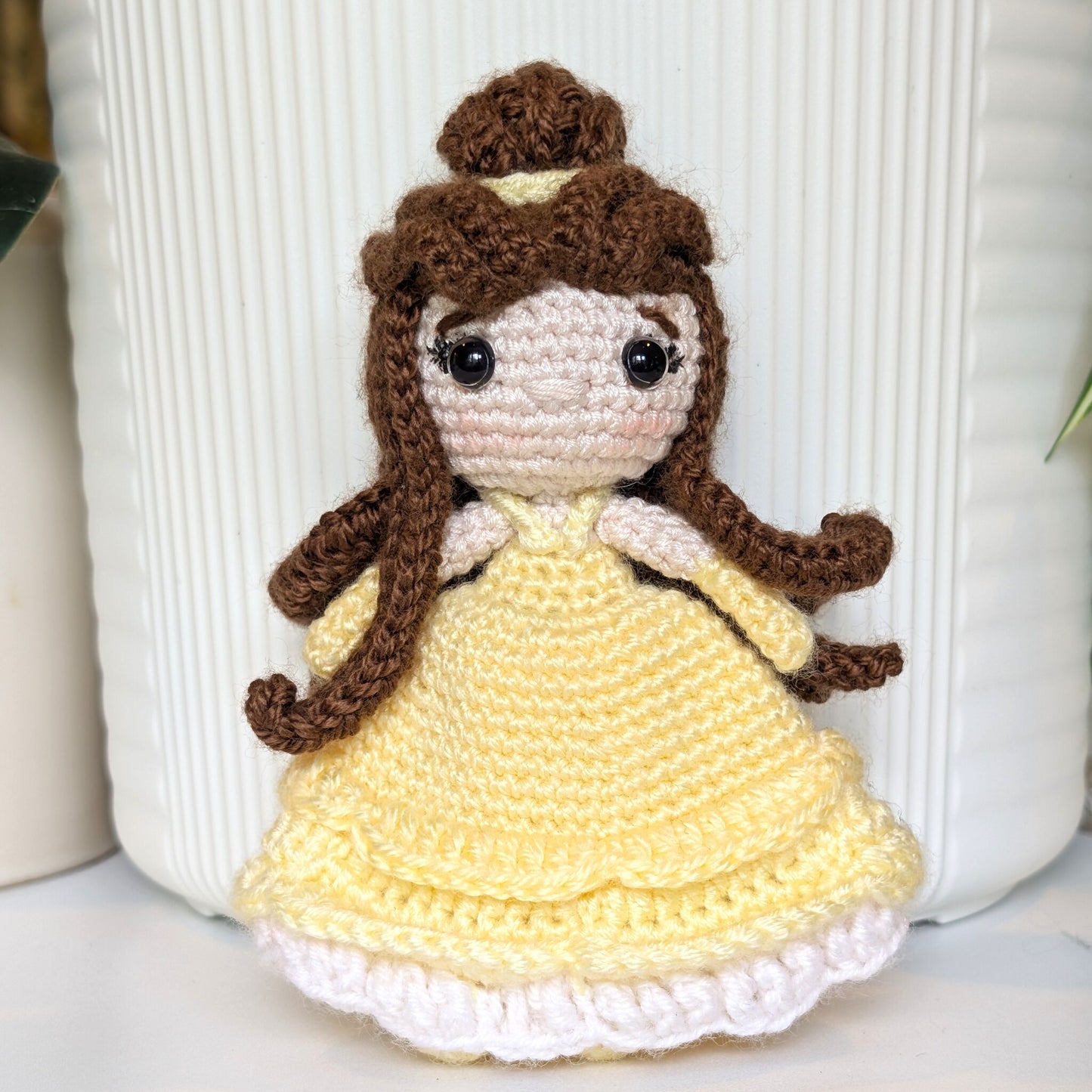 A crocheted doll with long brown hair, a cream-colored face, and a yellow dress with a white ruffled hem, standing in front of a white ribbed container.
