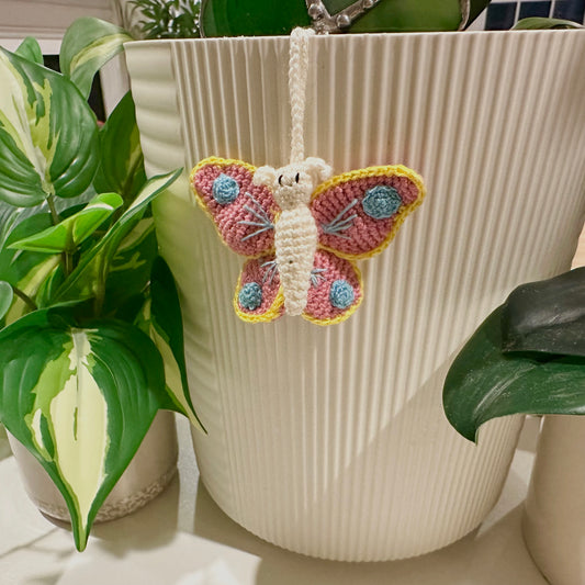 Crocheted butterfly decoration hanging from a plant pot with green plants in the background.