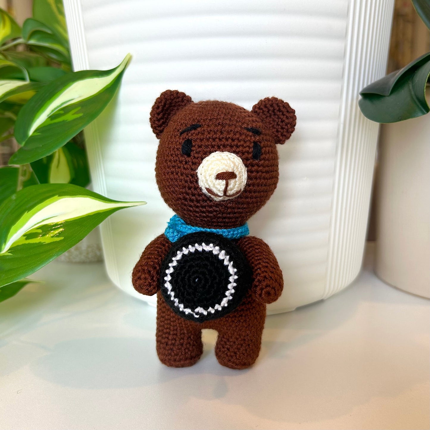 Brown crocheted bear with an azure scarf standing and holding a black ball with a white stripe