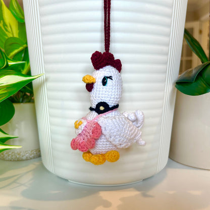 White crocheted chicken charm wearing a salmon and light pink apron, and a black flower around the neck