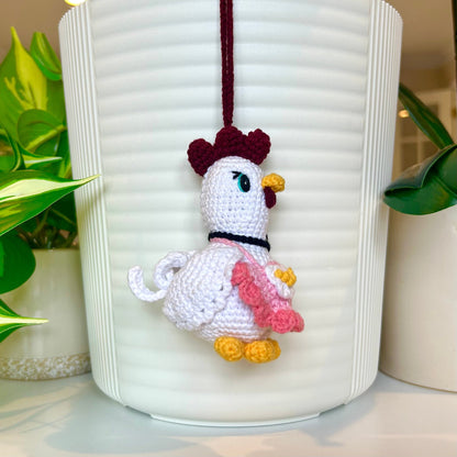 Left side profile of White crocheted chicken charm wearing a salmon and light pink apron, and a black flower around the neck