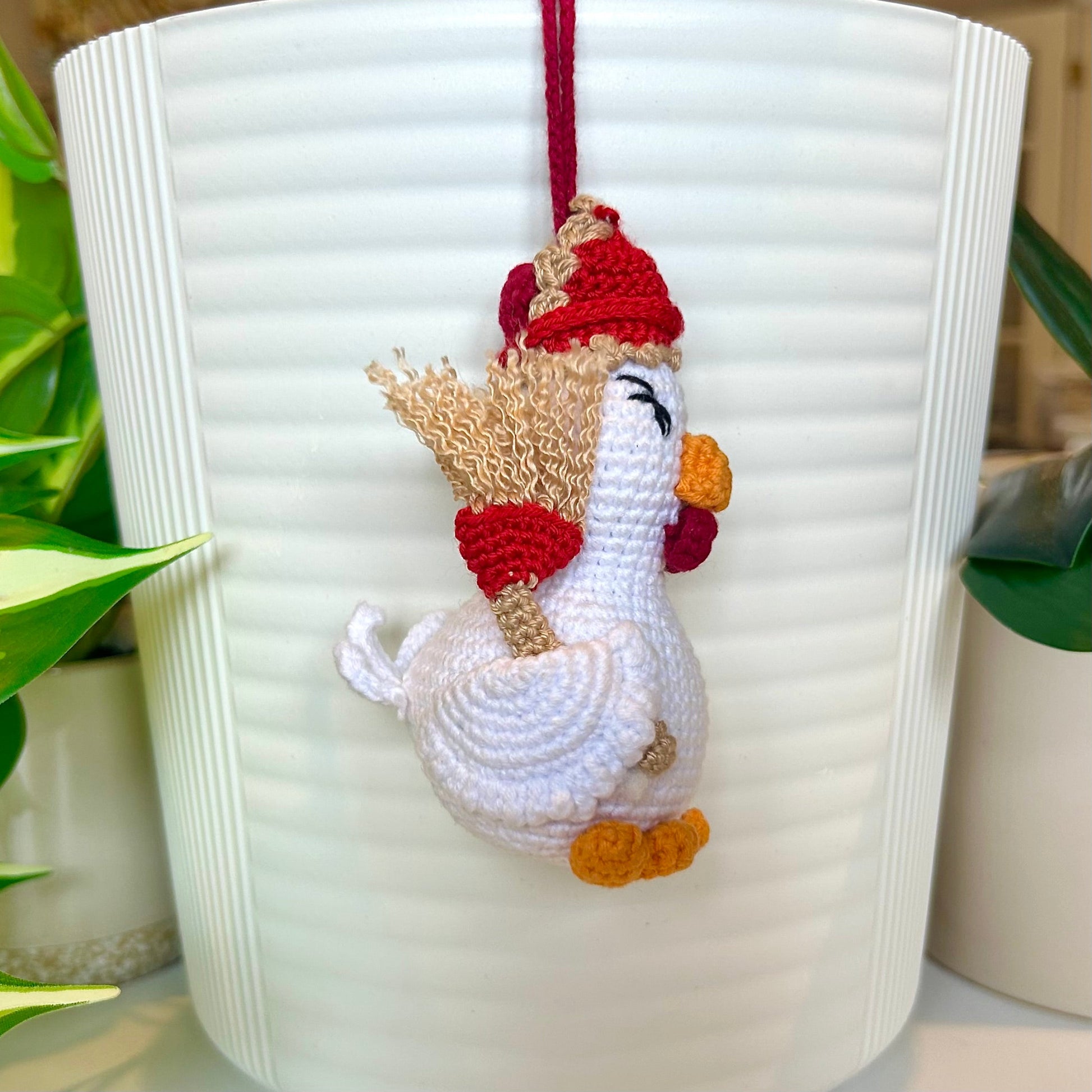 Left side profile of white crocheted chicken charm wearing a red and tan hat and carrying a broom