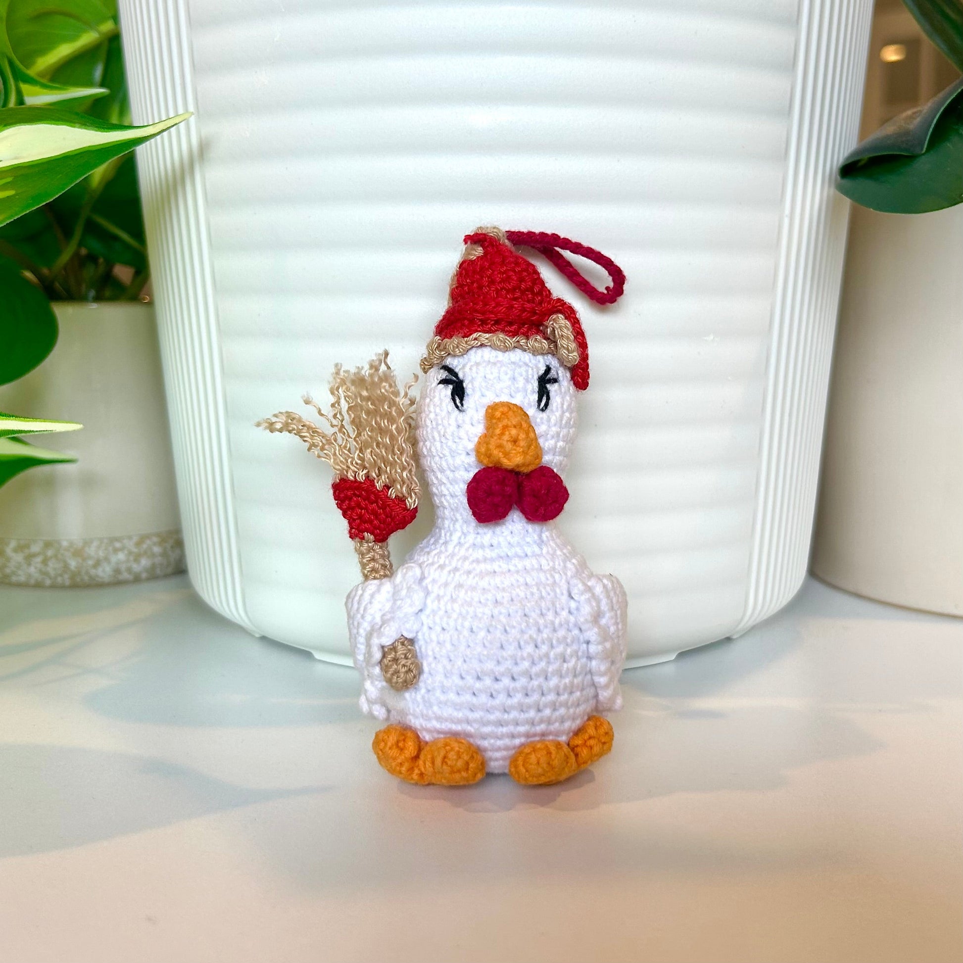 White crocheted chicken charm wearing a red and tan hat and carrying a broom
