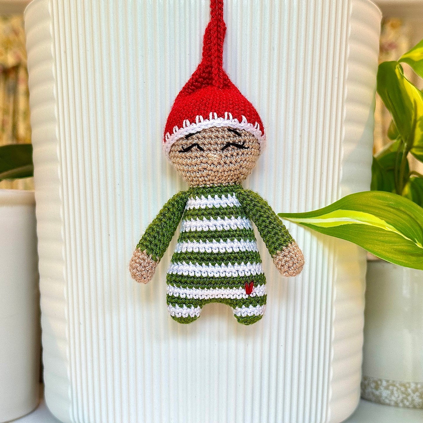 A crocheted hanging elf ornament in moss green outfit with a red hat, almond beige face, squinting, displayed in front of a white ribbed vase