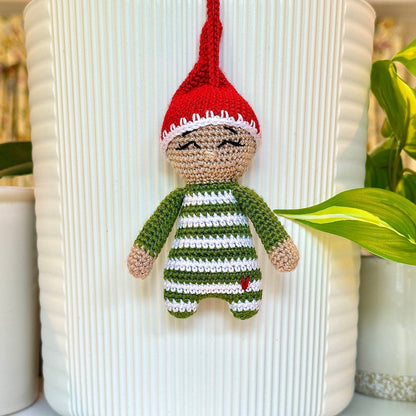 A crocheted hanging elf ornament in moss green outfit with a red hat, almond beige face, squinting, displayed in front of a white ribbed vase