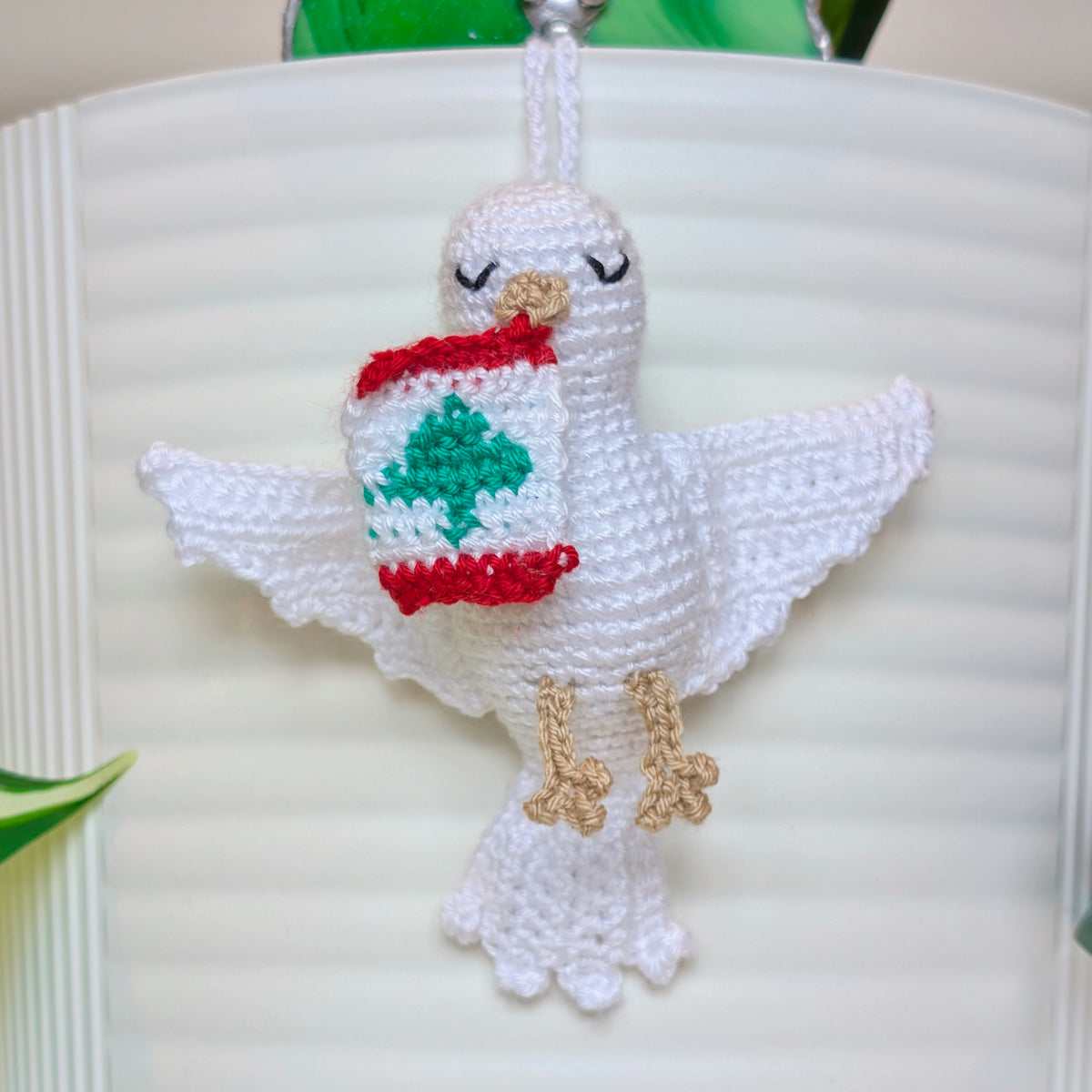 A white crocheted dove ornament with closed eyes, holding a Lebanon flag in beak, hanging against a white ribbed background.