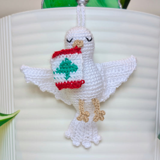 A white crocheted dove ornament with closed eyes, holding a Lebanon flag in beak, hanging against a white ribbed background.