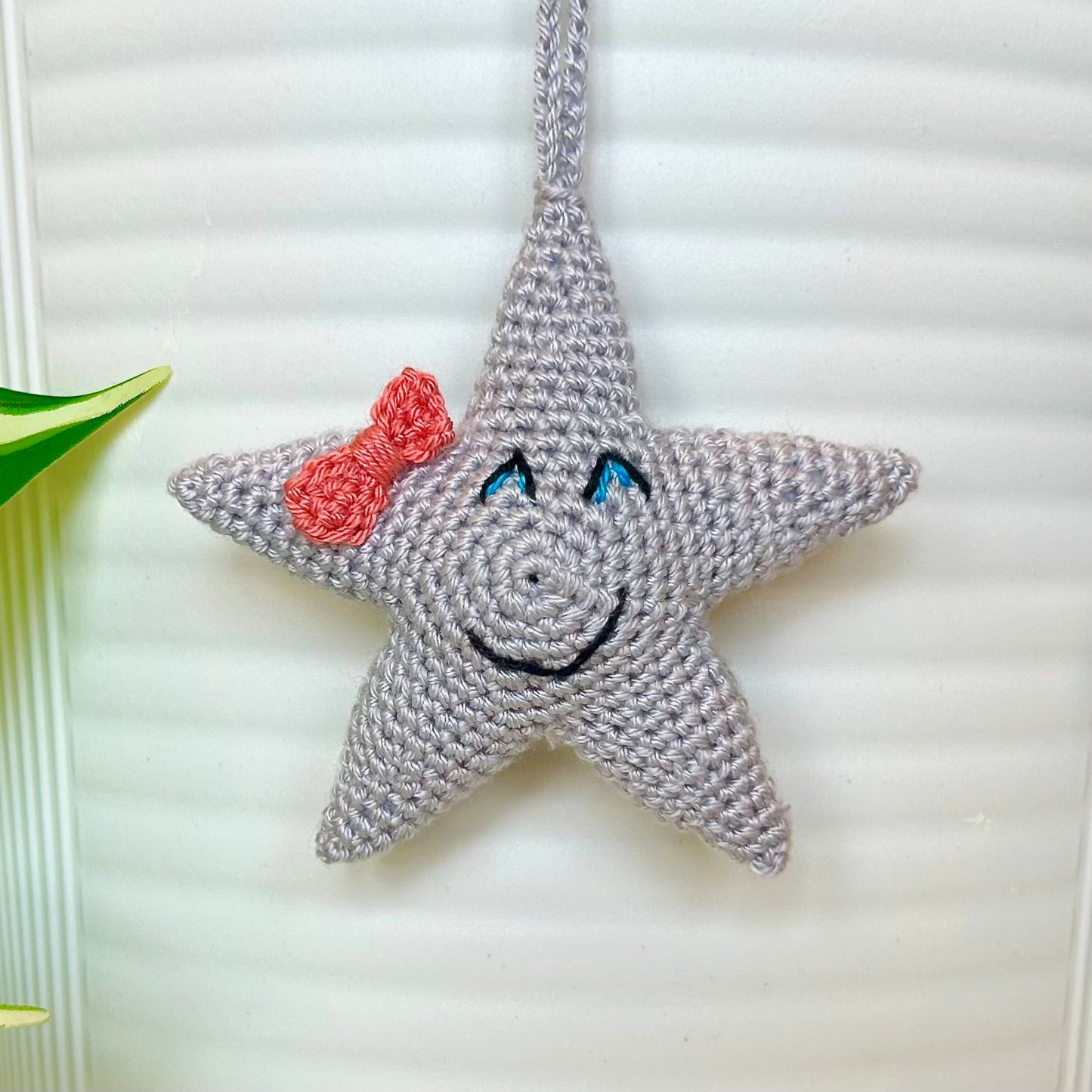 Gray crocheted star charm with a coral pink bow, blue eyes, and a smile