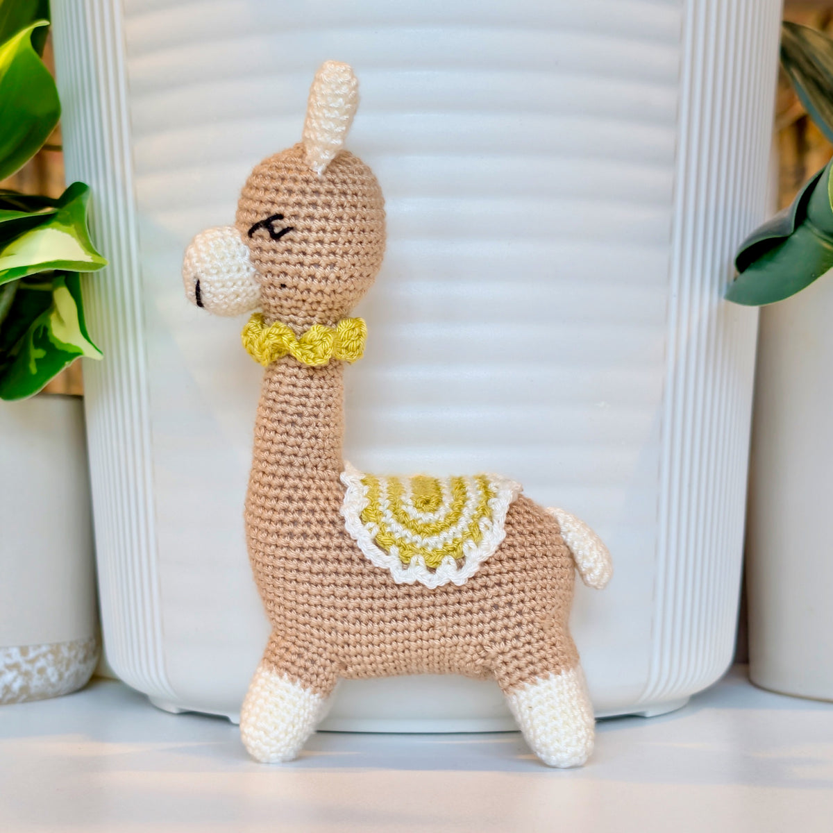Side view of crocheted vicuna llama beige plush with a white and chartreuse saddle and a chartreuse necklace