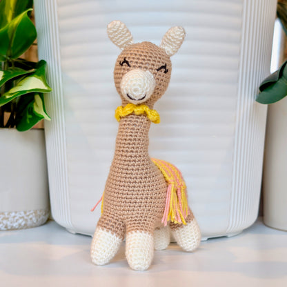 Crocheted vicuna llama beige plush with a yellow and pink saddle and a yellow necklace